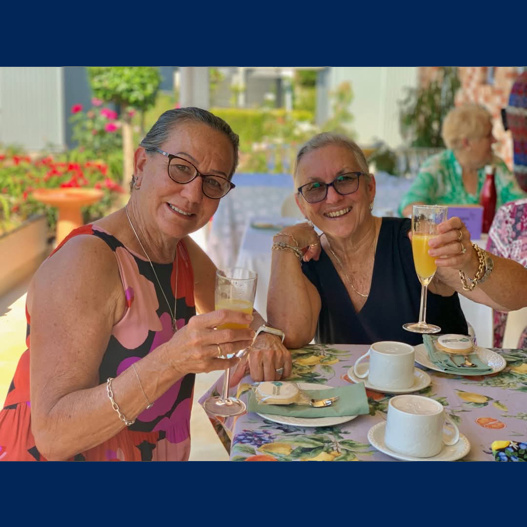 Two women sitting at an outside table, each with a drink in hand, looking at the camera and smiling