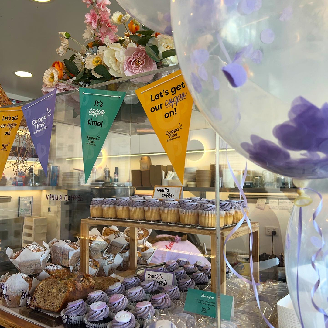 A bakery decorated for a Cuppatime for Dementia event. There's a glass display cabinet full of cupcakes and various sweets and Cuppatime bunting has been put up.