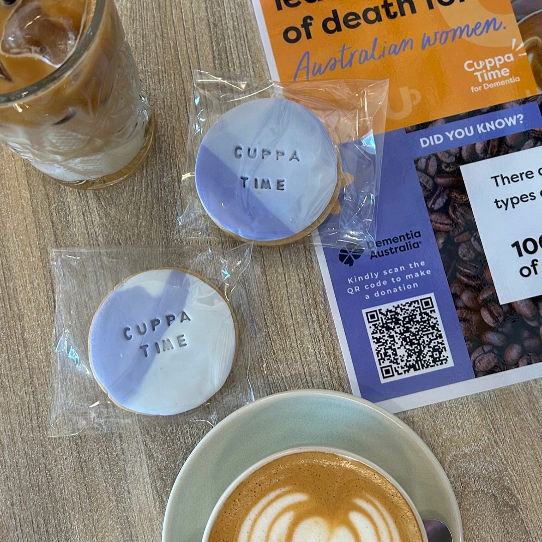A bird's eye view of a table with a coffee, a cold drink, two Cuppatime biscuits decorated with purple icing and a dementia pamphlet.