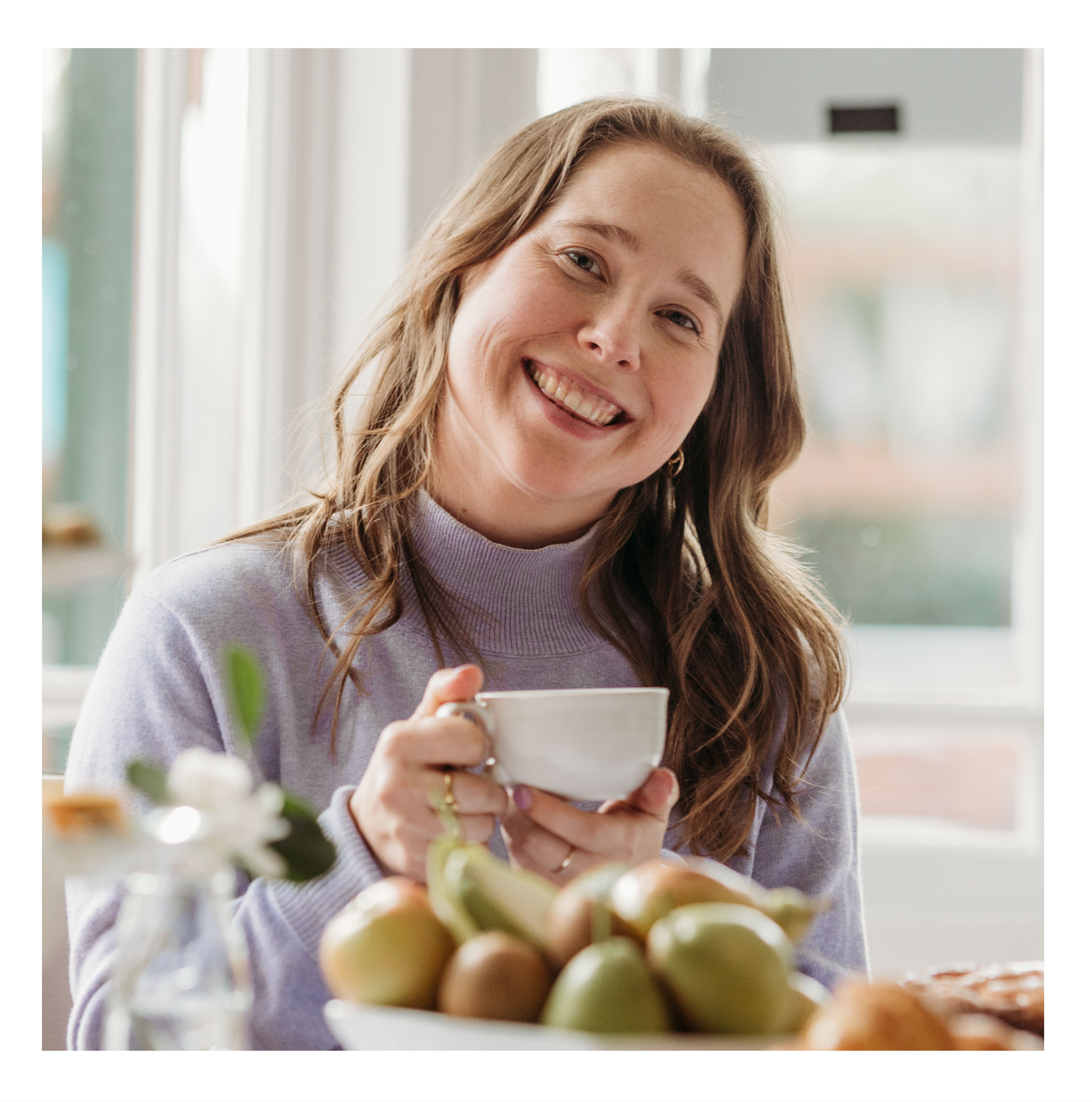 A young woman sitting down, holding a cup of tea with both hands, wearing a lavender sweater, looking at the camera and smiling.